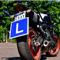 Motocykl z tablicą 'L' na parkingu, widok z tyłu, pomarańczowe felgi, tablica rejestracyjna DSW.