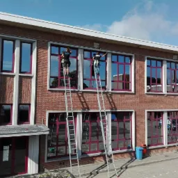 Dwóch pracowników na drabinach myje okna na wysokości w budynku z czerwonej cegły z fioletowymi ramami okiennymi.
