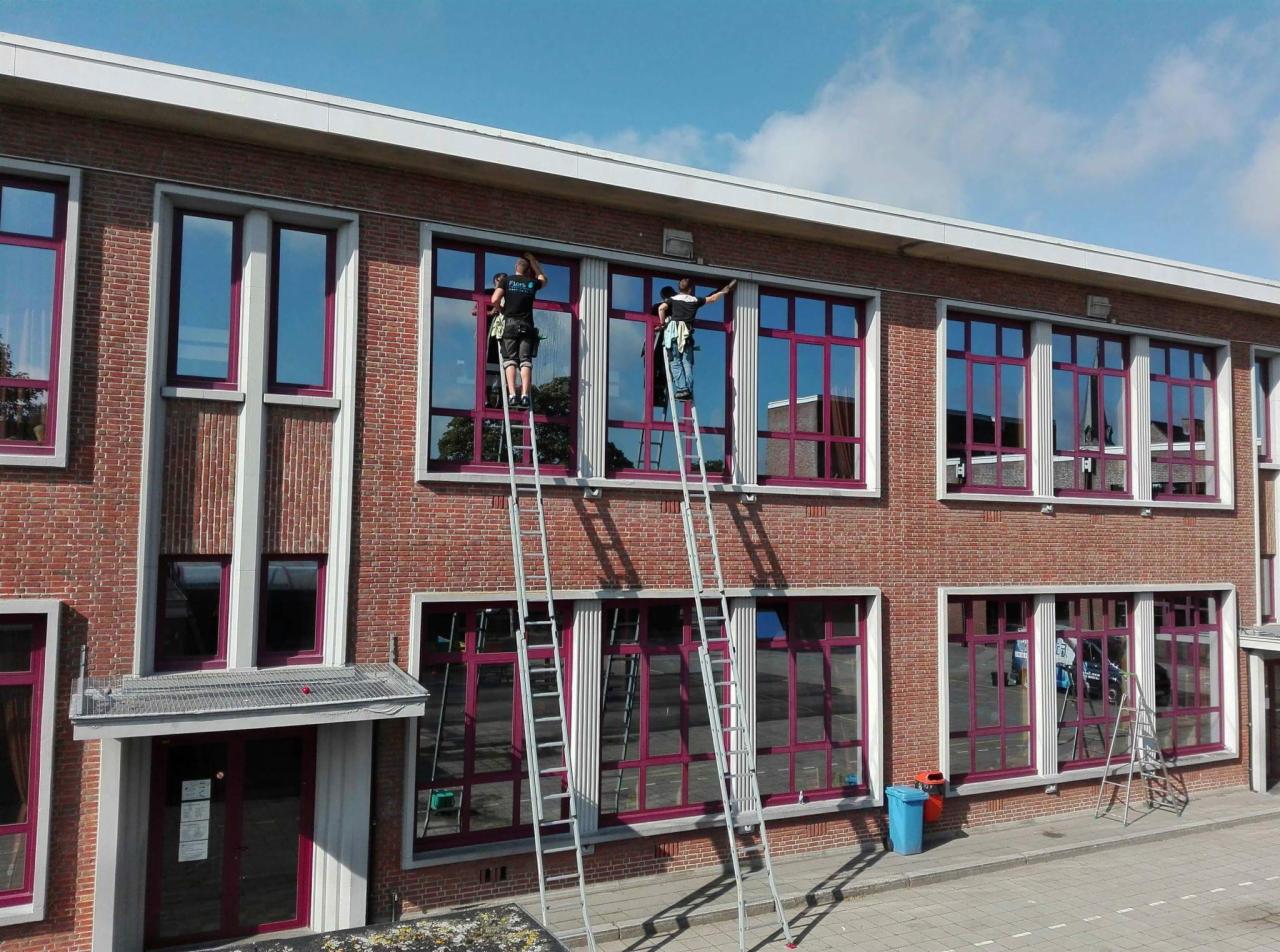 Dwóch pracowników na drabinach myje okna na wysokości w budynku z czerwonej cegły z fioletowymi ramami okiennymi.