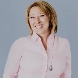 Portret uśmiechniętej kobiety w różowej koszuli na tle szarego tła, z widocznym srebrnym naszyjnikiem w kształcie łzy.