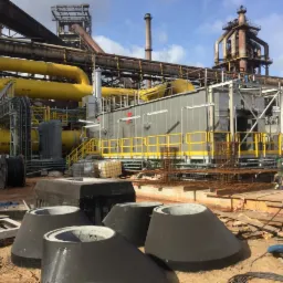 Konstrukcja przemysłowa z żółtymi rurami, stalowymi elementami i betonowymi prefabrykatami w kształcie ściętych stożków na pierwszym planie, teren budowy.