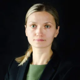 Portret kobiety z blond włosami spiętymi w kucyk, ubranej w czarną marynarkę i zieloną sukienkę, na czarnym tle.