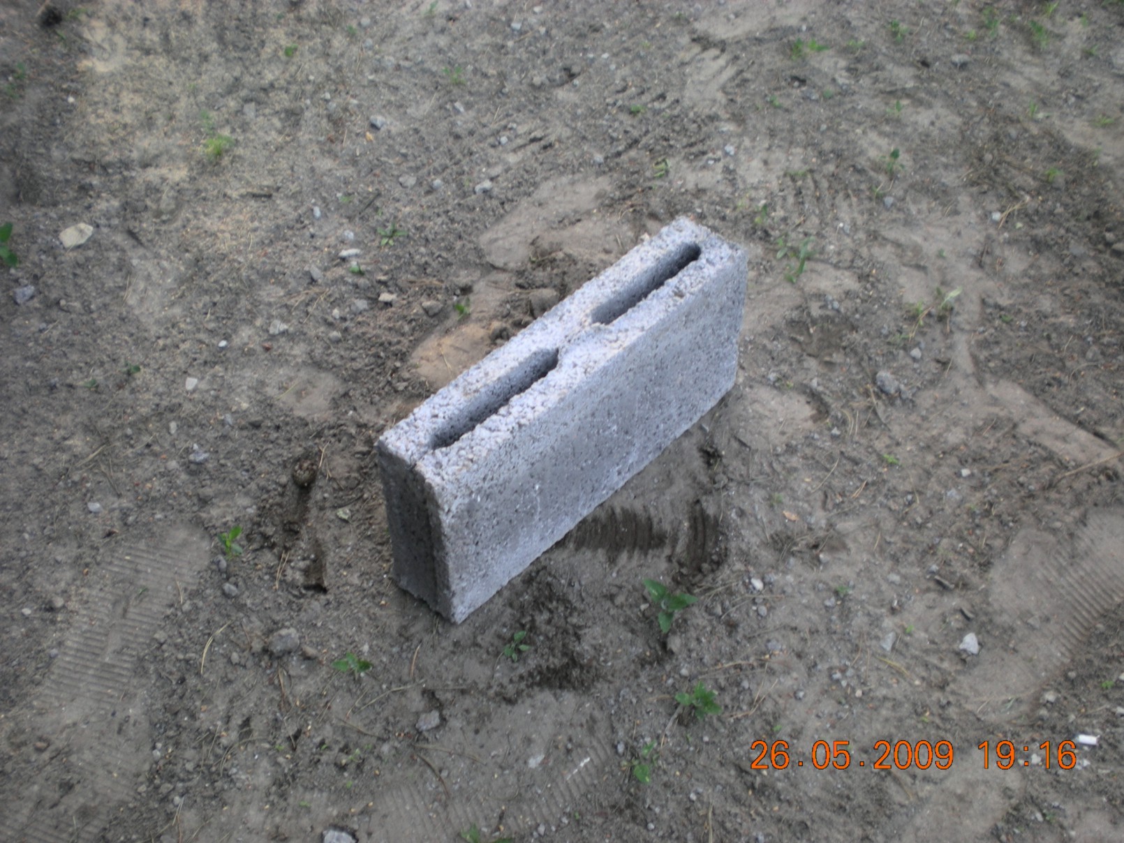 Pustak betonowy z dwoma otworami leżący na ziemi z widocznymi śladami opon i dacie wykonania zdjęcia.