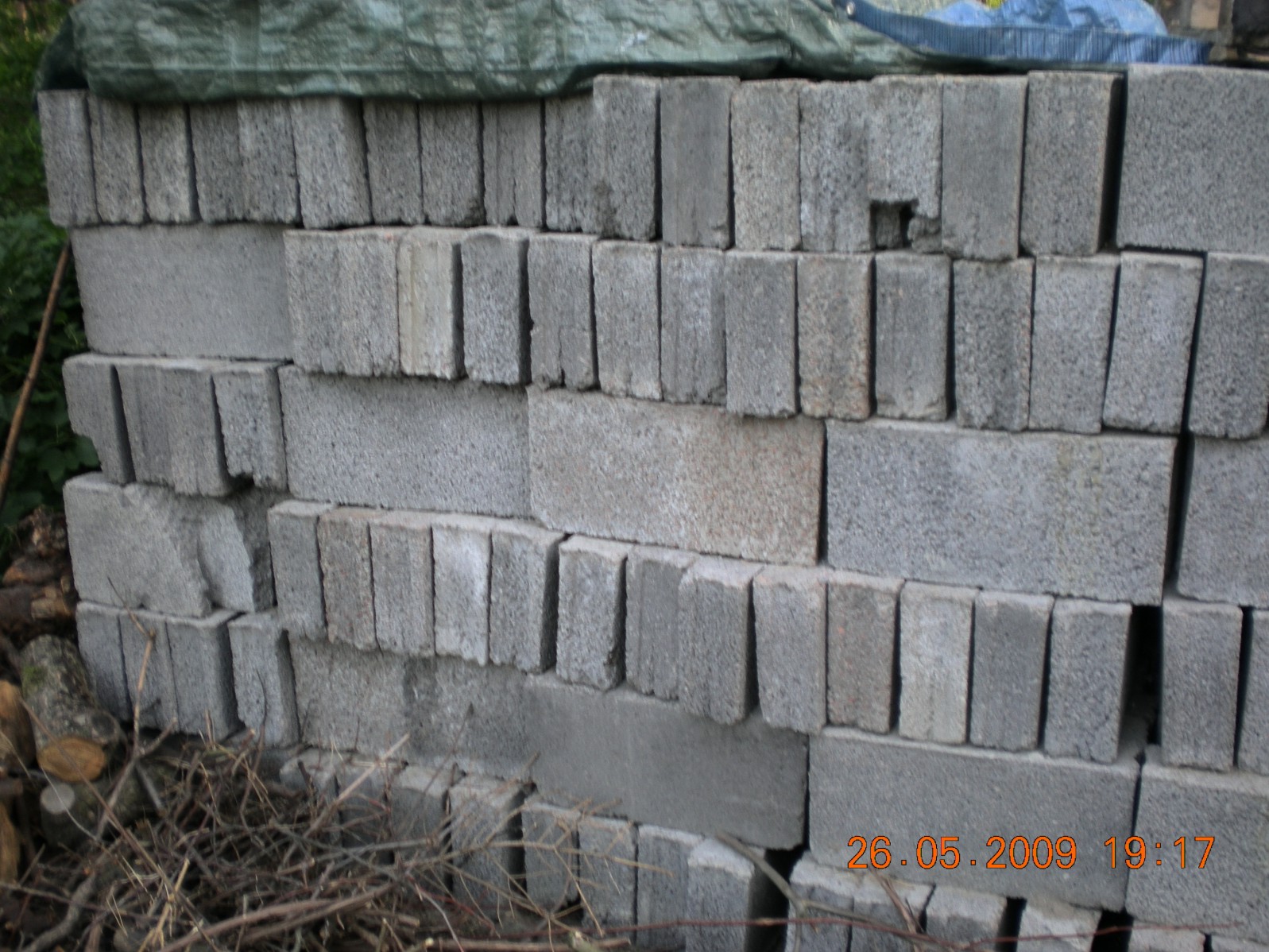 Stos szarych bloczków betonowych ułożonych warstwami, częściowo przykrytych zieloną plandeką, z widocznymi gałęziami w dolnej części kadru.