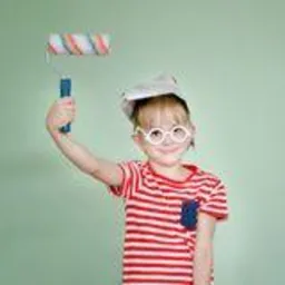 Uśmiechnięta dziewczynka w okularach i czapce maluje ścianę wałkiem z kolorowymi paskami. Na ścianie widoczne świeże ślady farby.