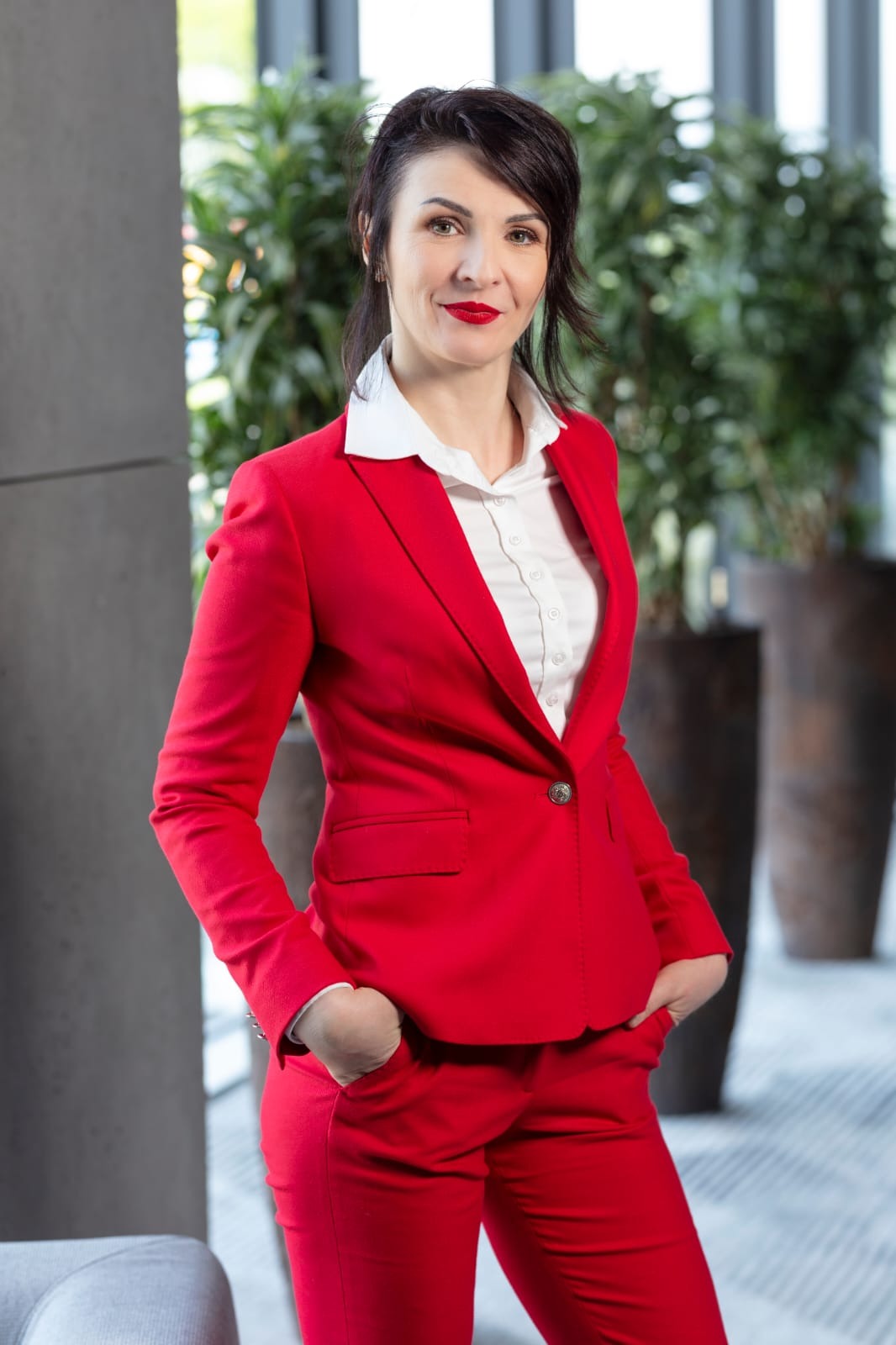 Uśmiechnięta kobieta w czerwonym garniturze i białej koszuli, stojąca na tle roślin doniczkowych, sugerująca profesjonalizm w branży finansowej.