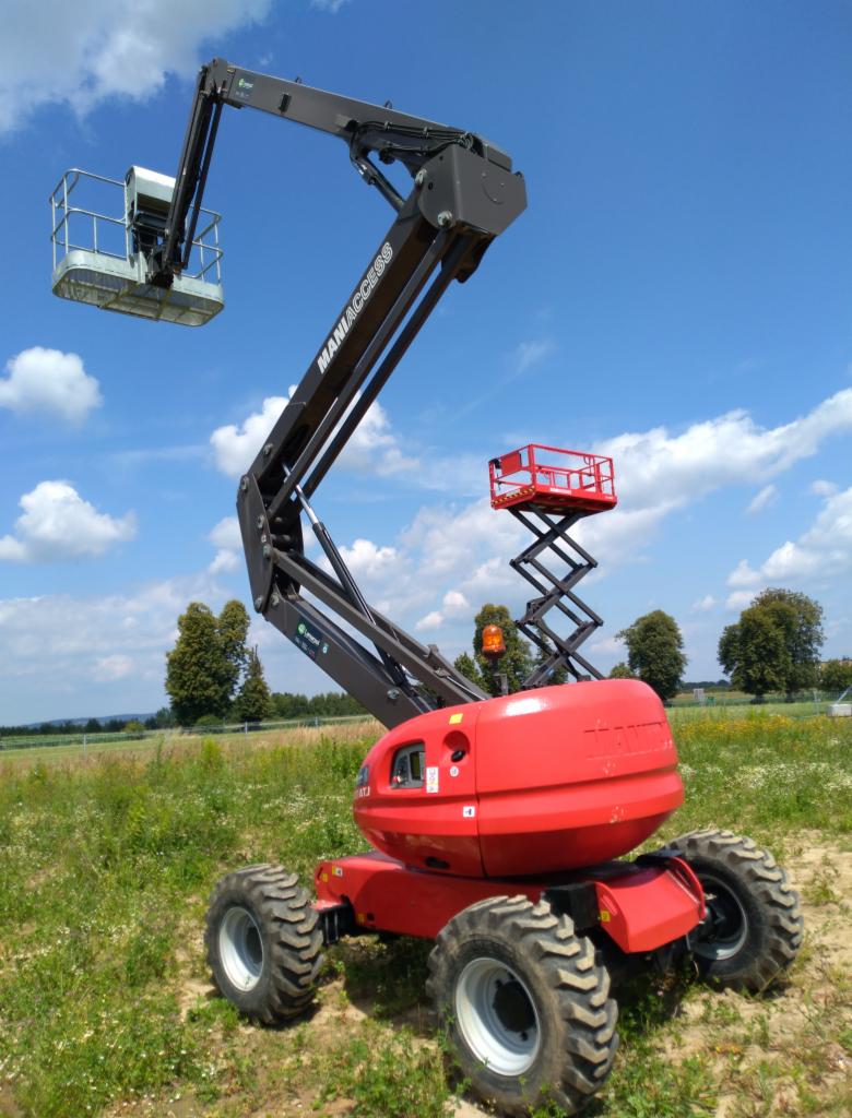 Czerwony wózek podnośnikowy teleskopowy Maniac Access z wysięgnikiem i koszem na tle błękitnego nieba z chmurami i zielonego pola. W tle widoczny drugi, mniejszy podnośnik nożycowy.