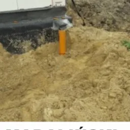 Wkopana pomarańczowa rura kanalizacyjna wystająca z ziemi przy ścianie budynku w trakcie budowy. Widoczna czarna folia hydroizolacyjna.