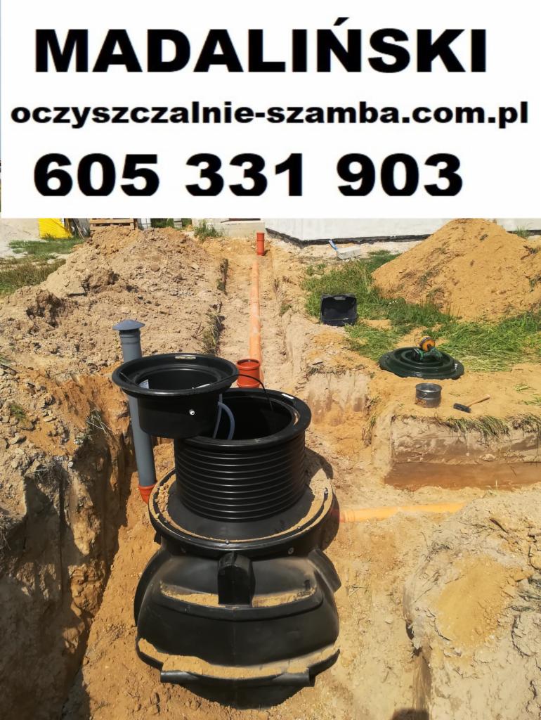 Montaż czarnej, plastikowej oczyszczalni ścieków w wykopie, widoczne rury kanalizacyjne i ziemia, w tle sterta piasku i trawa.