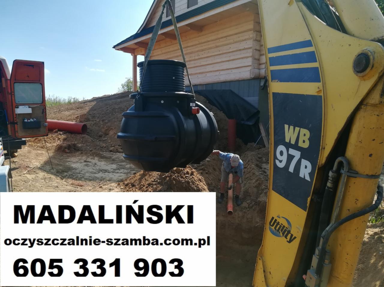 Żółta koparka opuszcza czarny zbiornik z tworzywa sztucznego do wykopu, w tle drewniany dom i pomarańczowe rury, widoczny pracownik w wykopie. Na pierwszym planie reklama firmy z numerem telefonu...