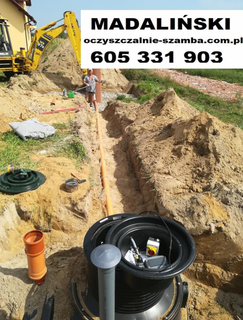 Montaż przydomowej oczyszczalni ścieków: wykop z rurą drenażową, widoczna koparka i pracownik z poziomicą, w tle dom jednorodzinny.