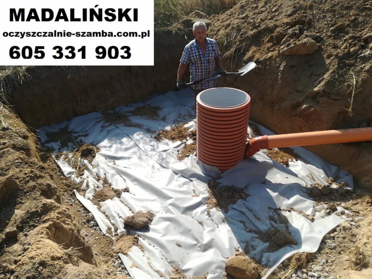 Wykop z wyłożoną geowłókniną, wewnątrz pionowy, pomarańczowy zbiornik z karbowanej rury i podłączony do niego poziomy rurociąg. W tle mężczyzna z łopatą. Na górze zdjęcia widoczny baner reklamowy...