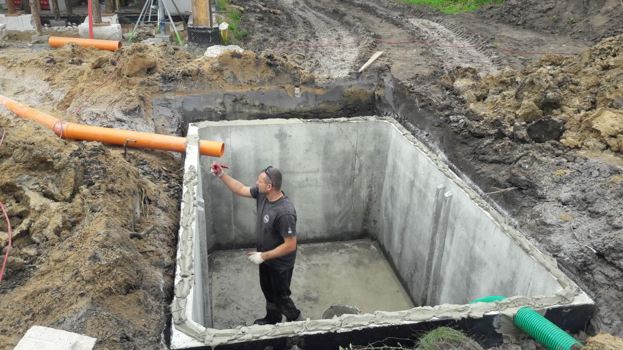 Pracownik uszczelnia betonowy zbiornik w wykopie, widoczne rury kanalizacyjne wchodzące do zbiornika, ziemia wokół wykopu, częściowo błotnista.
