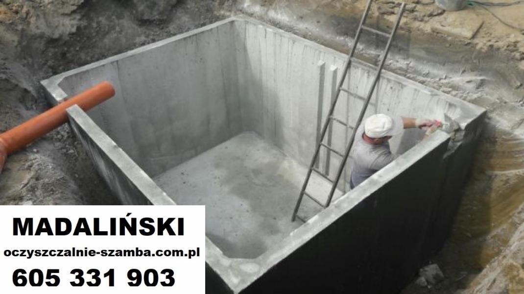 Betonowy zbiornik w wykopie, pracownik w kasku wygładza krawędzie, widoczna drabina i rura kanalizacyjna.