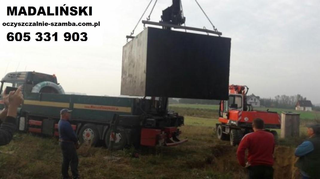 Ciężki, betonowy zbiornik opuszczany przez dźwig na plac budowy, widoczny transport ciężarowy i osoby nadzorujące montaż, w tle zabudowa jednorodzinna.