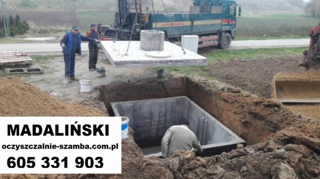 Montaż betonowego zbiornika: robotnicy umieszczają prefabrykowany element w wykopie za pomocą dźwigu, widoczny wykop, palety i ciężarówka w tle.