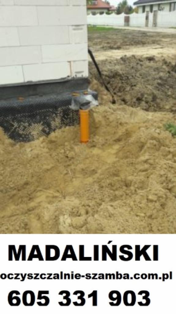 Wkopana pomarańczowa rura kanalizacyjna wystająca z ziemi przy ścianie budynku w trakcie budowy. Widoczna czarna folia hydroizolacyjna.