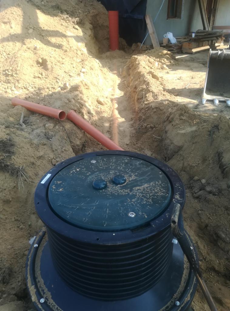 Montaż szamba betonowego z zieloną pokrywą w wykopie, widoczne rury kanalizacyjne i fragment budynku w tle.