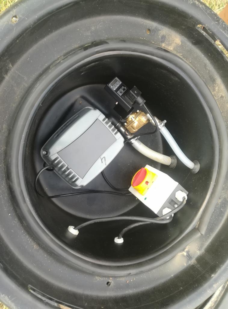 Wnętrze czarnego zbiornika z zamontowanym sterownikiem, elektrozaworem i przewodami doprowadzającymi wodę. Widoczny panel sterujący z szarą obudową oraz przycisk awaryjny koloru czerwonego.