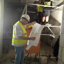 Trzech elektryków w kamizelkach odblaskowych i kaskach pracuje na rusztowaniu, jeden z nich analizuje plan instalacji, widoczne kable i metalowe korytka kablowe pod sufitem.