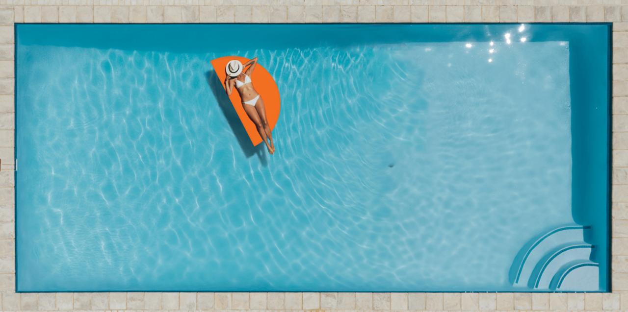 Kobieta w białym bikini i kapeluszu leży na pomarańczowym materacu w turkusowym basenie z kamiennymi brzegami, widok z góry.