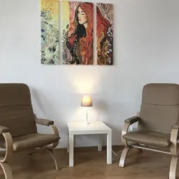 Dwa beżowe fotele relaksacyjne z drewnianą ramą ustawione naprzeciwko siebie, pomiędzy nimi biały stolik z lampką, na ścianie tryptyk z portretem kobiety o rudych włosach.