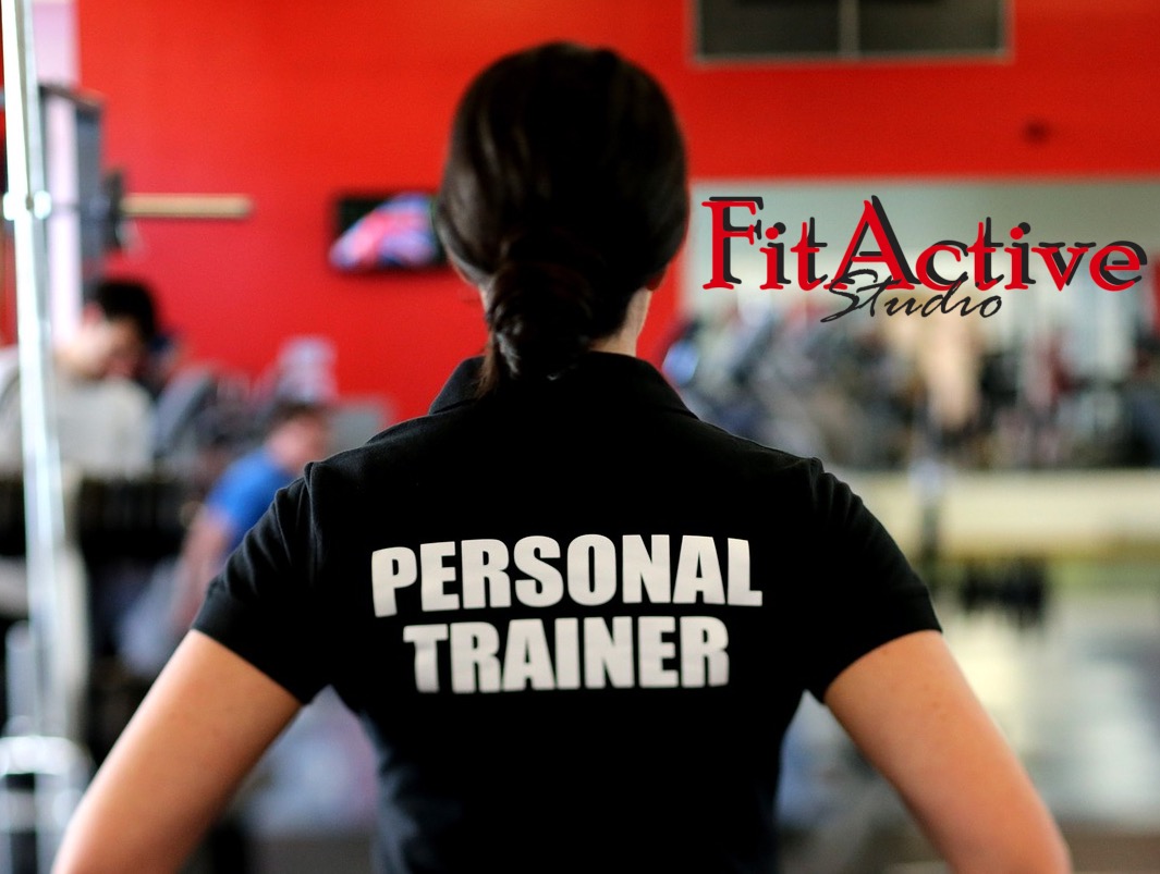 Kobieta tyłem w czarnej koszulce z napisem 'PERSONAL TRAINER' na tle siłowni z czerwonymi ścianami i logiem 'FitActive Studio' na szklanej ścianie.