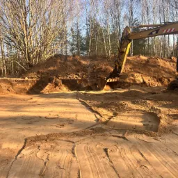 Żółta koparka CAT podczas prac ziemnych na piaszczystym terenie, widoczne ślady opon i wykopany rów w ziemi, w tle drzewa bez liści.