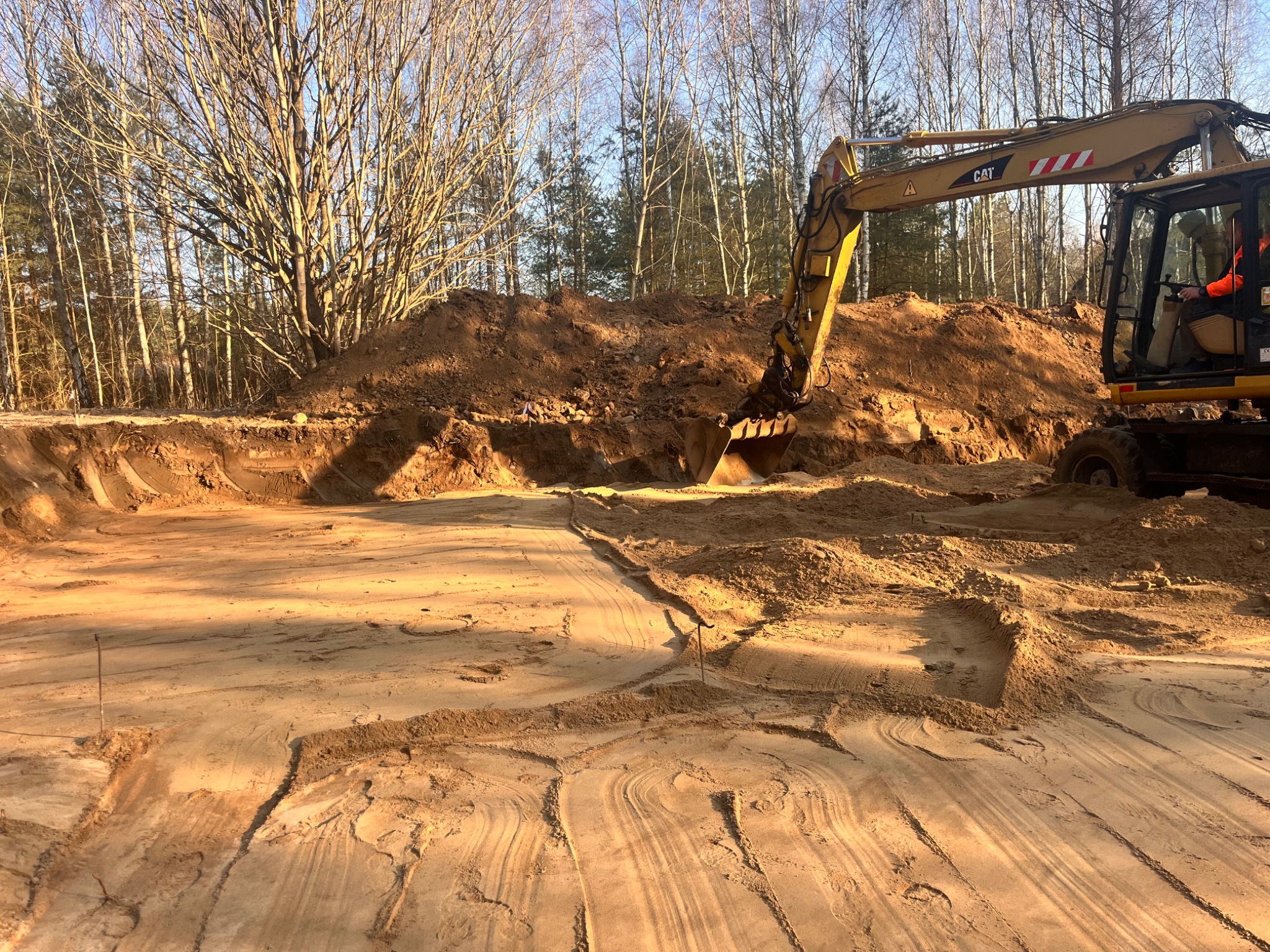 Żółta koparka CAT podczas prac ziemnych na piaszczystym terenie, widoczne ślady opon i wykopany rów w ziemi, w tle drzewa bez liści.