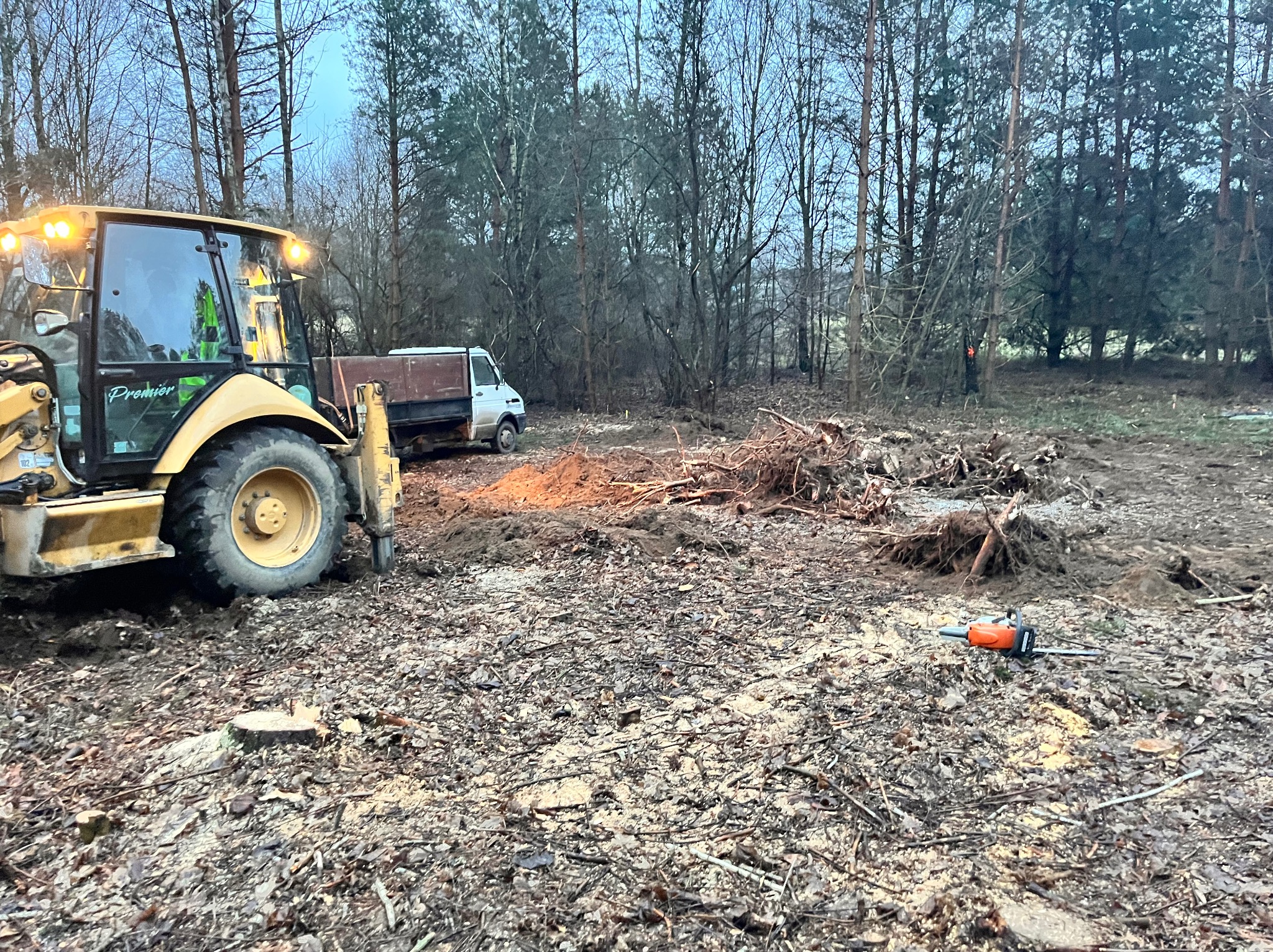 Żółta koparko-ładowarka z włączonymi światłami stoi na terenie po wycince drzew, obok leży piła łańcuchowa, a w tle widoczny jest samochód dostawczy i las.