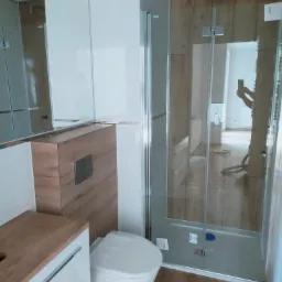 Nowoczesna łazienka z kabiną prysznicową walk-in, drewnopodobnymi płytkami na ścianach i podłodze, białą ceramiką i szafką podumywalkową z drewnianym blatem. W lustrze odbija się osoba robiąca...