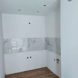 Fragment wykończonego pomieszczenia z drewnopodobną podłogą, białymi ścianami, pasem płytek imitujących marmur oraz widoczną kratką wentylacyjną i instalacjami hydraulicznymi.