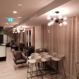 Nowoczesne oświetlenie sufitowe z żarówkami w stylu retro w przestronnym wnętrzu, widoczne krzesła barowe i fotele, perforowany sufit i znak ewakuacyjny.