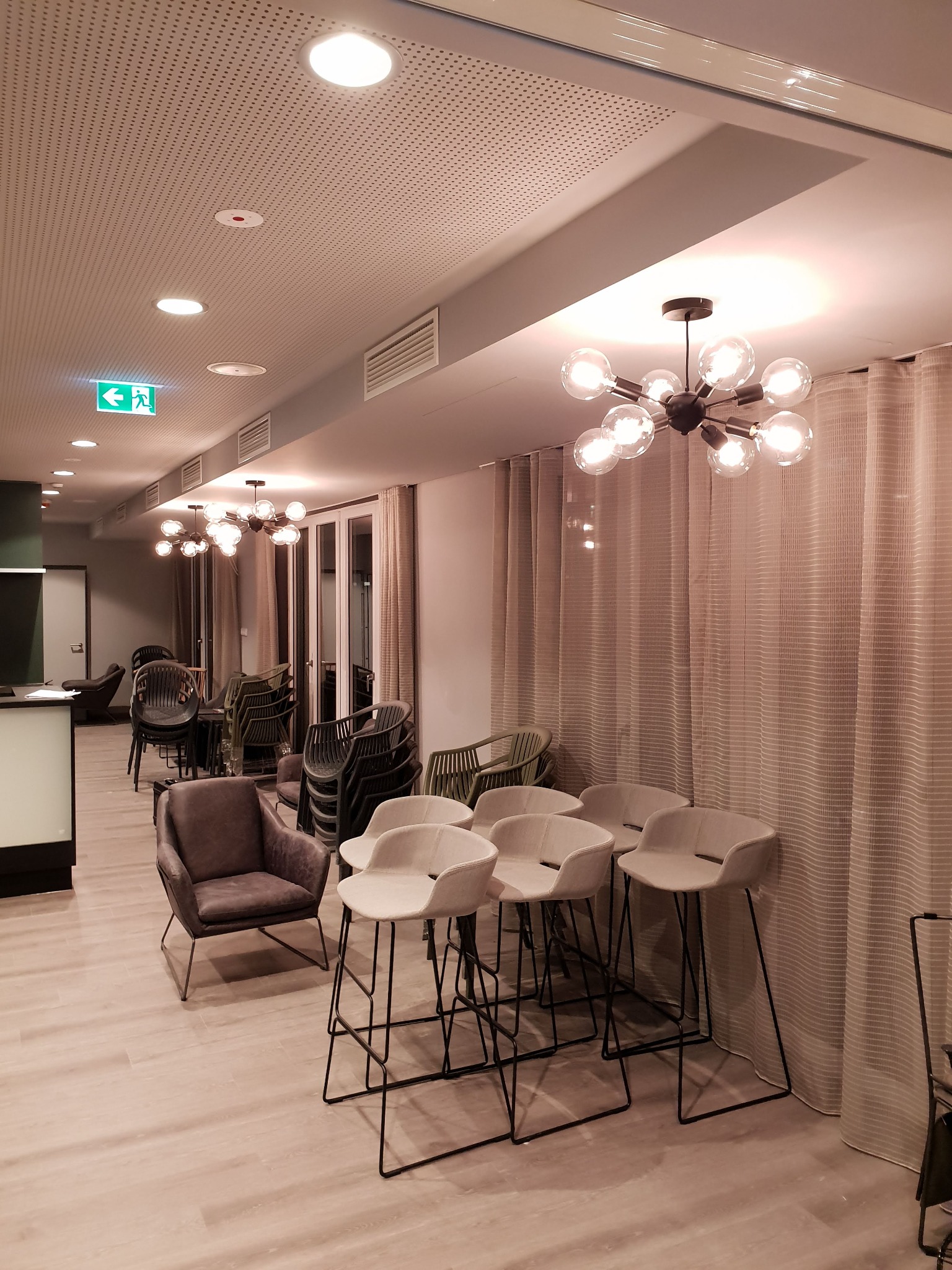 Nowoczesne oświetlenie sufitowe z żarówkami w stylu retro w przestronnym wnętrzu, widoczne krzesła barowe i fotele, perforowany sufit i znak ewakuacyjny.