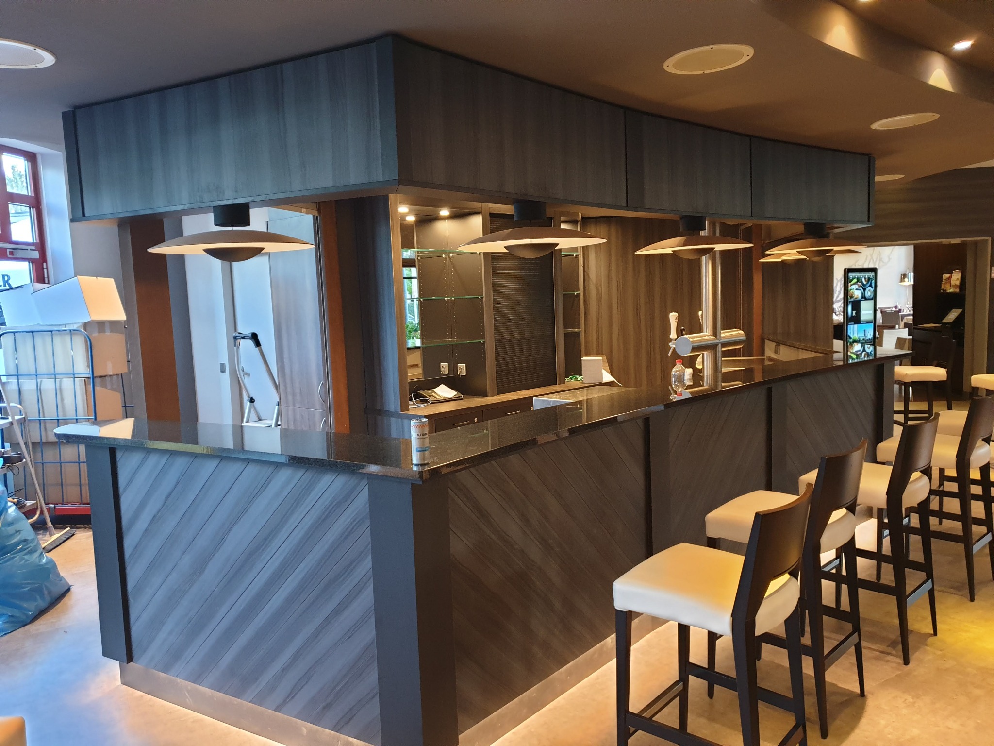 Nowoczesny bar z drewnianym wykończeniem, podświetlanym blatem i rzędem krzeseł barowych z jasną tapicerką, oświetlony wiszącymi lampami w kształcie talerzy.