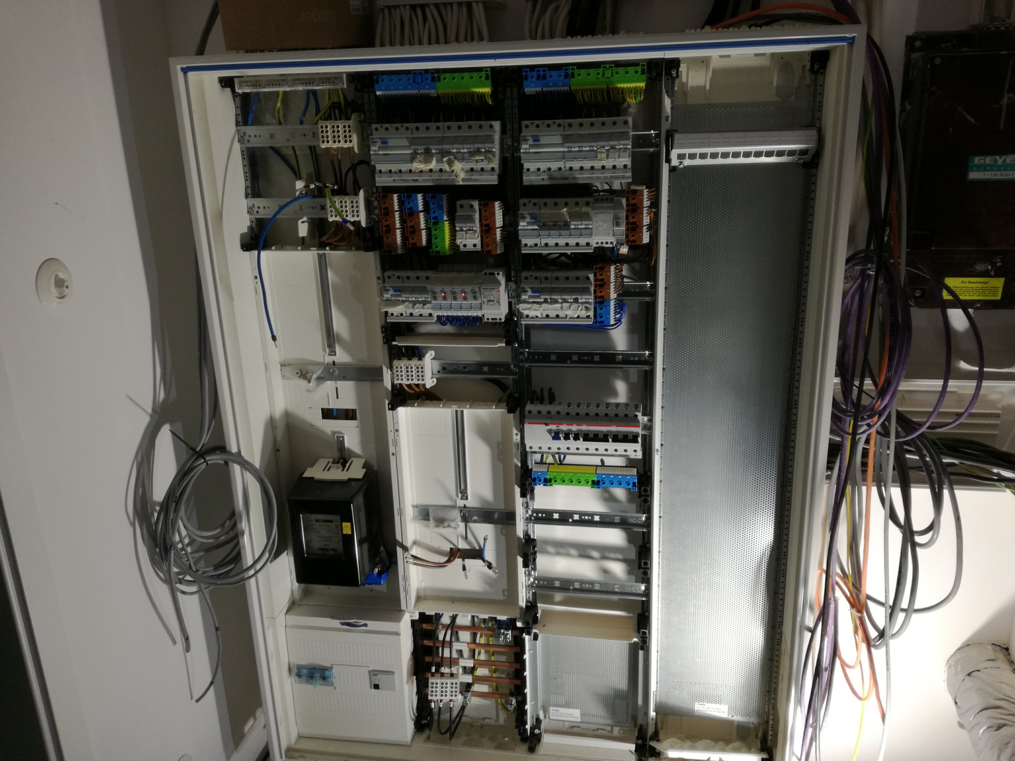 Otwarta skrzynka rozdzielcza z widocznymi bezpiecznikami, przewodami i licznikiem energii elektrycznej, zamontowana na białej ścianie, z widocznymi kablami wchodzącymi do skrzynki.