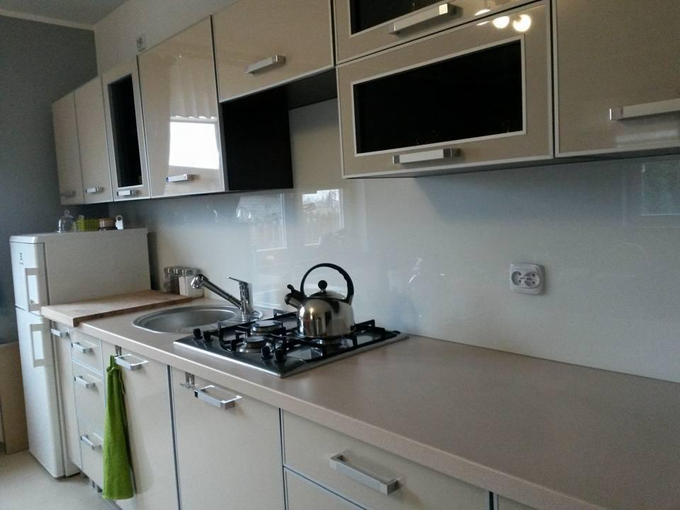 Jasna kuchnia z beżowymi szafkami, metalowymi uchwytami, szklanym panelem nad blatem, srebrnym czajnikiem na płycie gazowej i zieloną ścierką wiszącą na szafce.