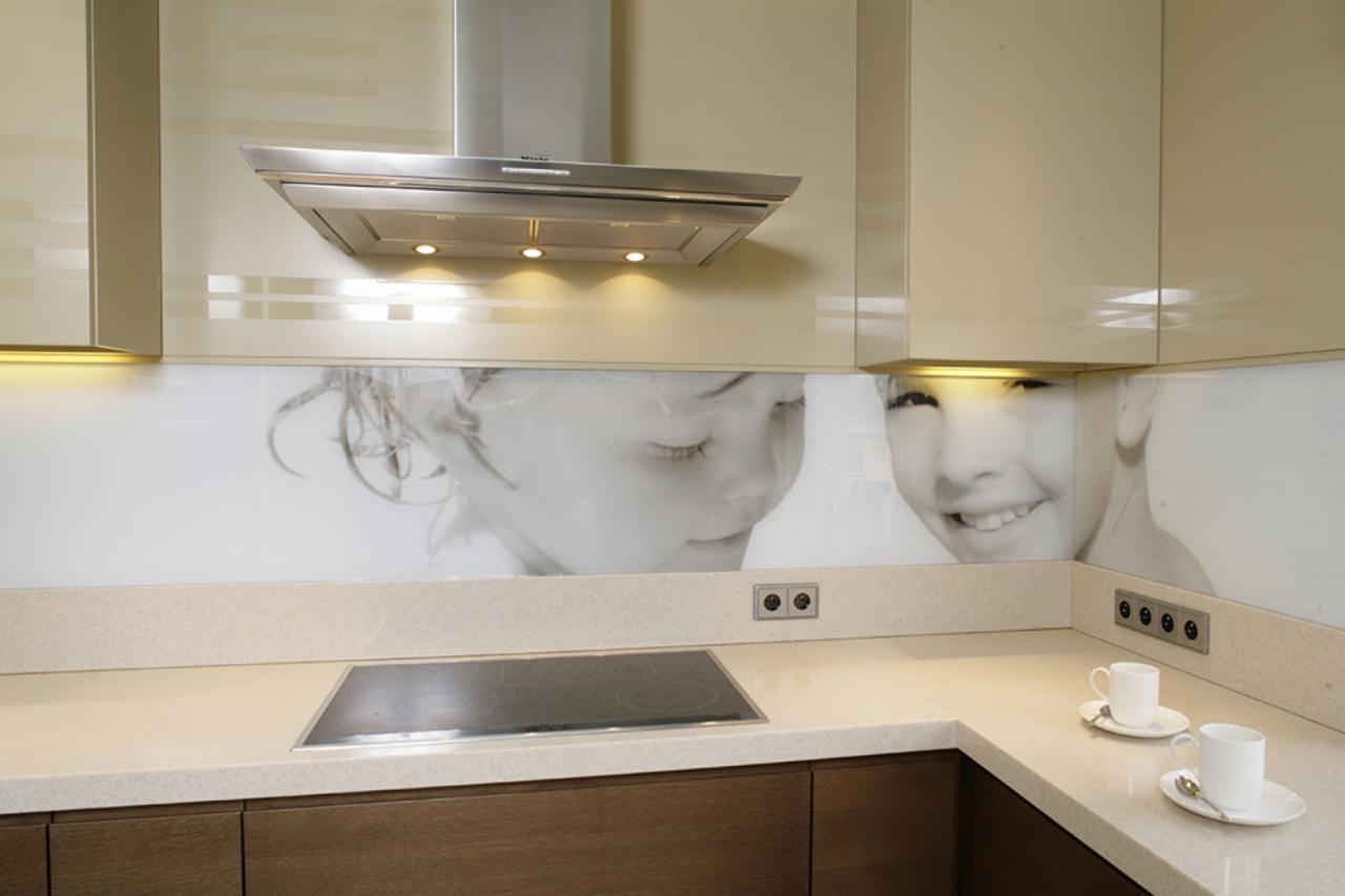 Nowoczesna kuchnia z kremowymi szafkami, szklanym panelem nad blatem z nadrukiem portretów dzieci, stalowym okapem i dwiema białymi filiżankami na blacie.