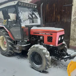 Czerwony traktor Zetor 7011 z pługiem do śniegu, częściowo pokryty śniegiem, stojący na ośnieżonym podwórku przed budynkiem z drewnianymi wrotami.