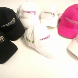 Kolekcja czapek z daszkiem w kolorach czarnym, białym i różowym, z wyhaftowanymi różnymi napisami i logo, ułożonych na białym tle.