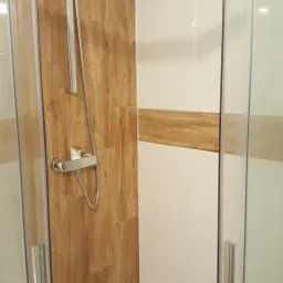 Kabina prysznicowa z przesuwnymi drzwiami, wykończona płytkami imitującymi drewno i białymi płytkami, z widoczną baterią i słuchawką prysznicową.