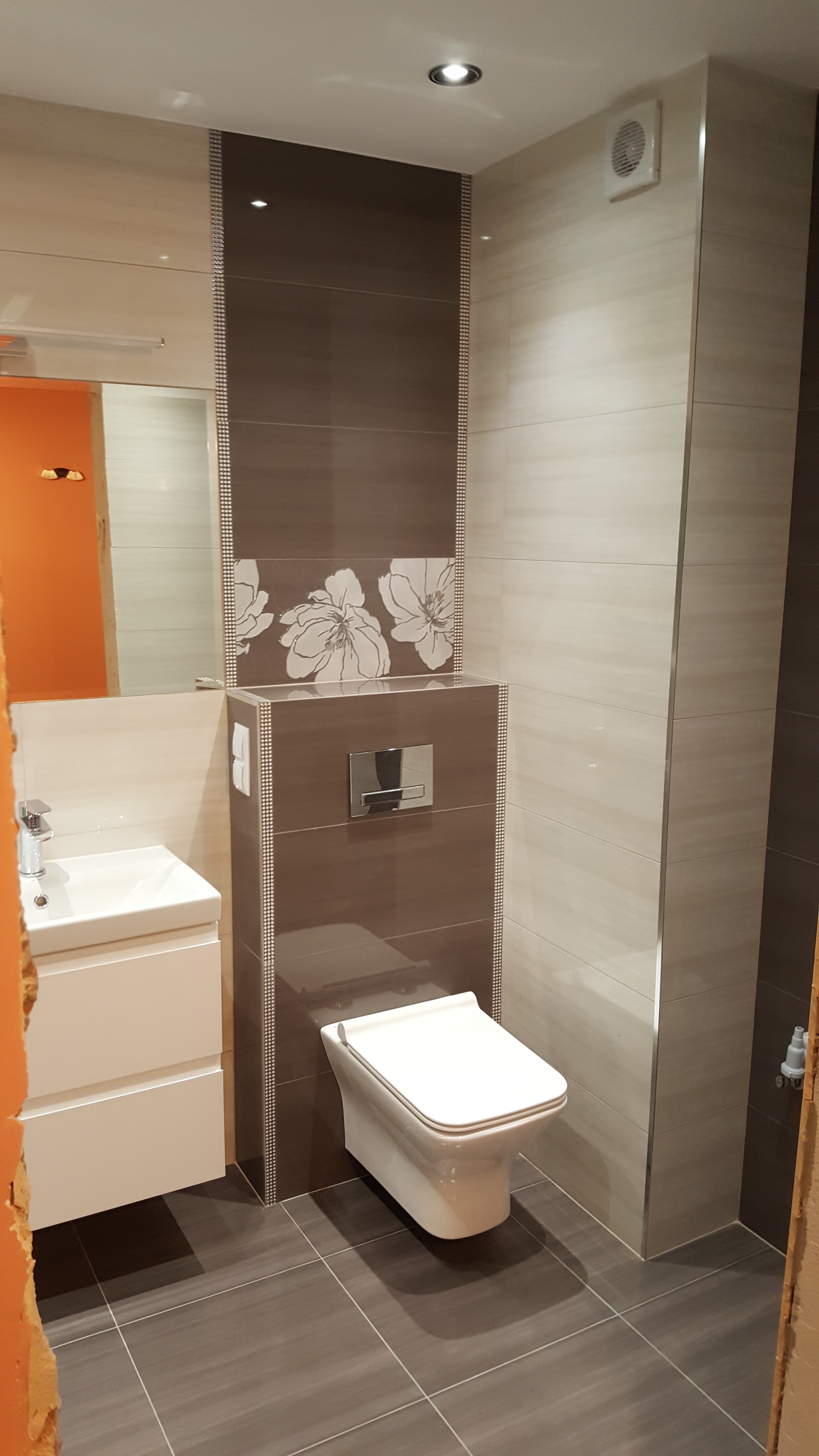 Wykończona łazienka z podwieszaną toaletą, umywalką z szafką, szaro-beżowymi płytkami z motywem kwiatowym i listwami dekoracyjnymi, widoczny fragment pomarańczowej ściany z sąsiedniego pomieszczenia.