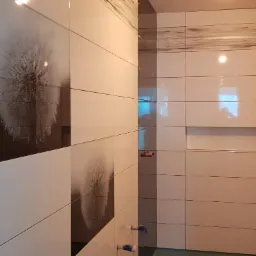 Ściana łazienki wyłożona białymi i ciemnoszarymi płytkami z motywem dmuchawca, widoczne krzyżyki dystansowe i wystające rury instalacyjne, oświetlenie punktowe w suficie.