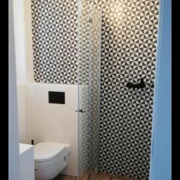 R-B TEAM - Nowoczesna łazienka z geometryczną czarno-białą mozaiką na ścianie, drewnopodobną podłogą, wiszącą toaletą, szklaną kabiną prysznicową i dekoracyjną lampą sufitową w stylu industrialnym.