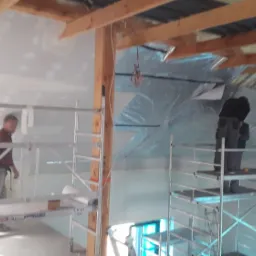 Prace wykończeniowe wewnątrz budynku: montaż izolacji pod dachem z użyciem rusztowań, widoczna drewniana konstrukcja więźby dachowej i częściowo wykończone ściany.