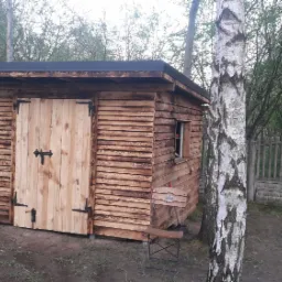 Drewniana szopa z czarnym dachem, wykonana z desek układanych poziomo, z pojedynczym oknem i drzwiami z metalowymi zawiasami, na tle brzozowego lasu i betonowego ogrodzenia.