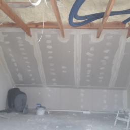 R-B TEAM - Poddasze w trakcie wykańczania: izolacja pianką na suficie, płyty gipsowo-kartonowe na ścianach, szpachlowanie łączeń. Widoczny pracownik przy ścianie, drabina i wiadro z zaprawą.