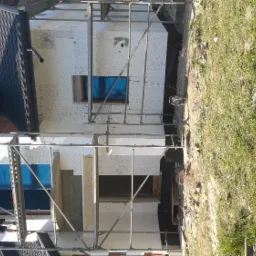 Dom w trakcie ocieplania elewacji, widoczne rusztowania, styropian na ścianach, okna zabezpieczone folią, pracownik na rusztowaniu.