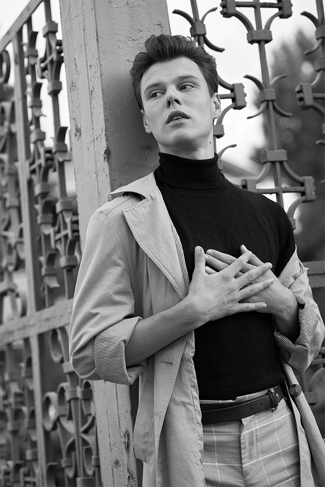 Młody mężczyzna w czarnym golfie i płaszczu, oparty o metalową bramę, patrzy w górę, ręce skrzyżowane na piersiach, zdjęcie czarno-białe.