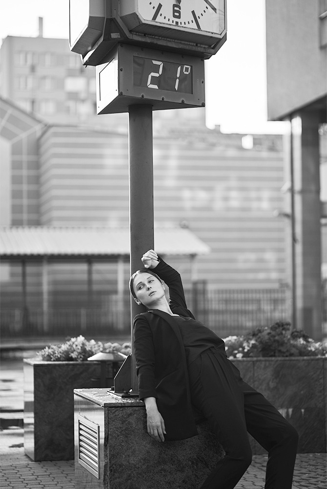 Czarno-białe zdjęcie kobiety w czarnym garniturze opierającej się o słup z zegarem i termometrem pokazującym 21 stopni, na tle miejskiego krajobrazu.